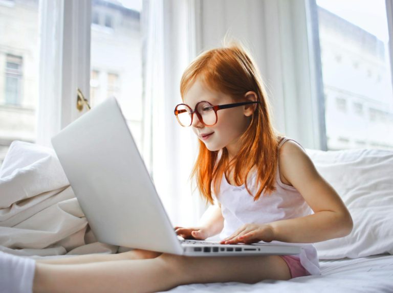 selective focus photo of young girl in glasses sitting in bed while using a laptop